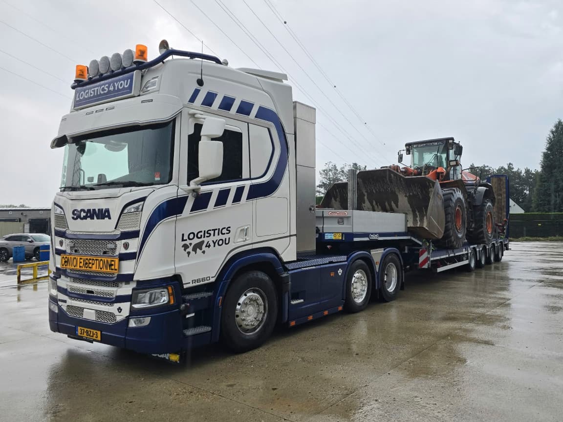 A Logistics4You Scania marked convoi exceptionnel hauling a wheel loader on a low-loader across a wet yard