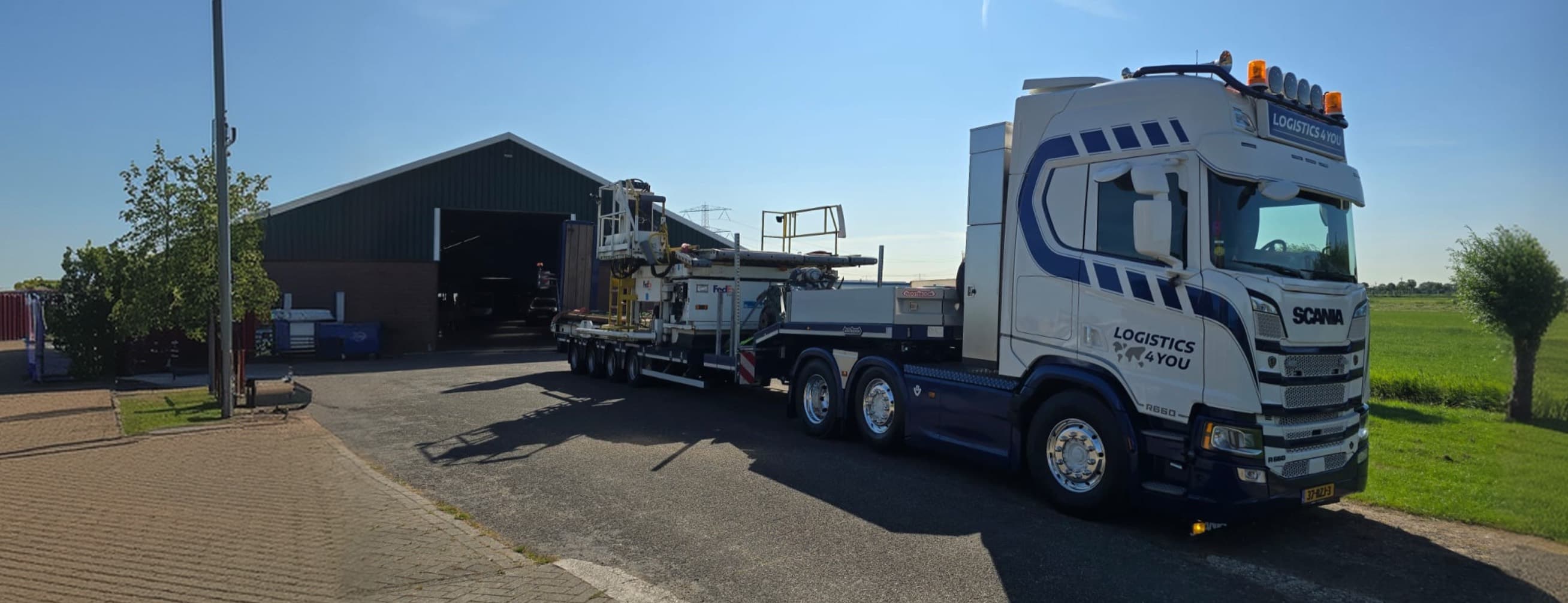 A Logistics4You Scania tractor unit with a loaded semi low-loader on a country road beside a green field